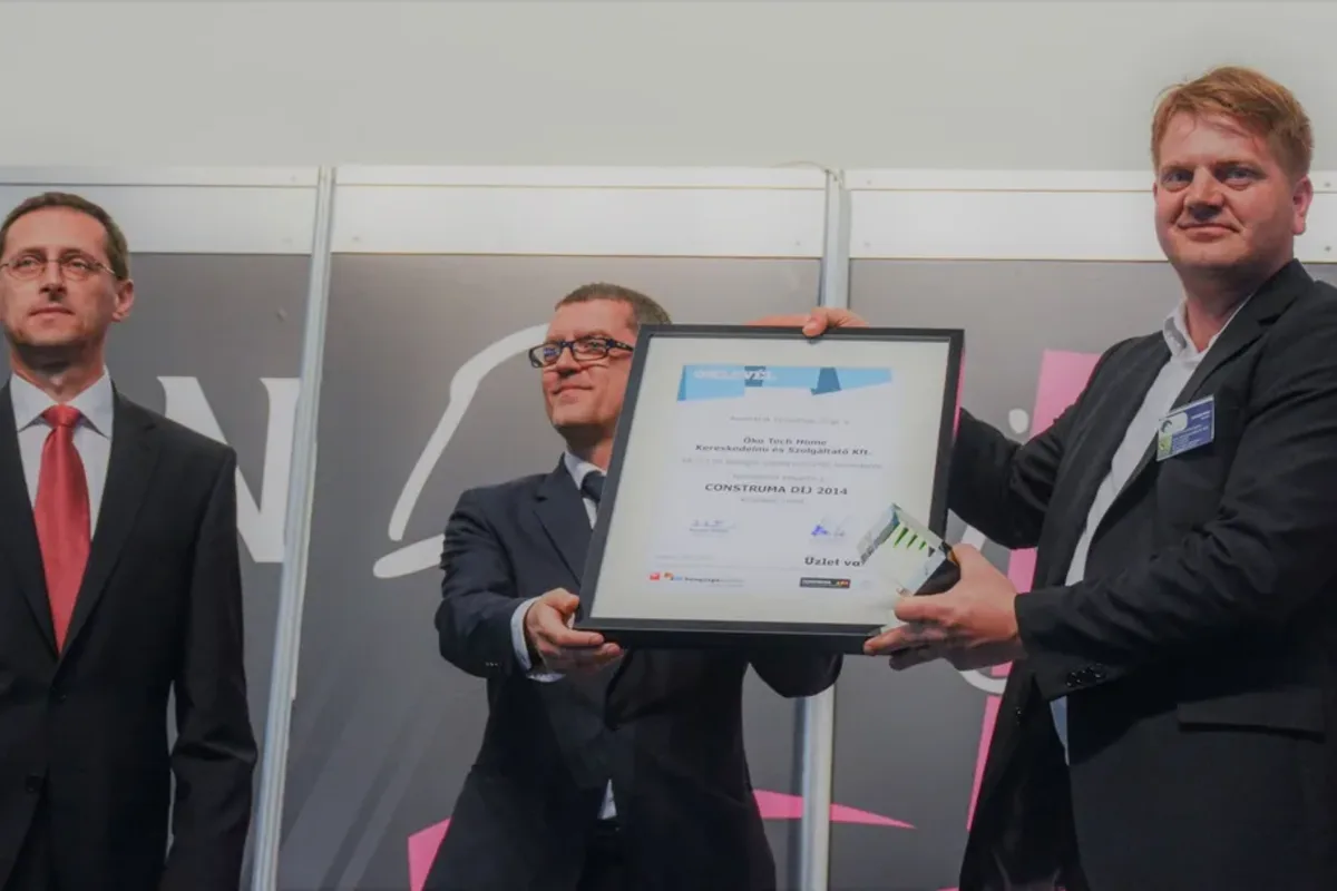 Keretezett oklevél átadása egy kiállítás színpadán: két öltönyös férfi tartja a „CONSTRUMA DÍJ 2014” feliratú oklevelet, mellettük egy harmadik férfi áll